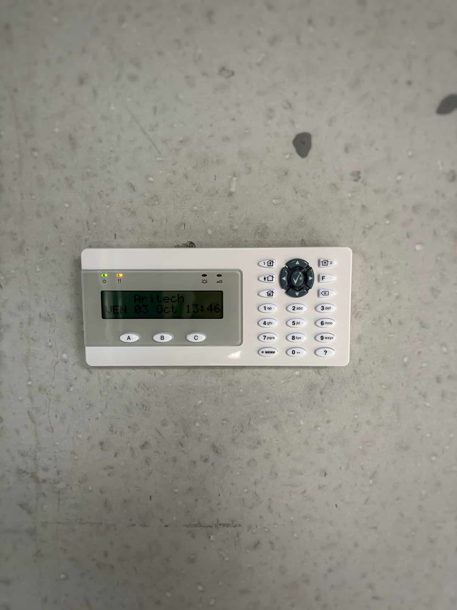 Clavier de centrale d'alarme Aritch sur mur gris affichant "VEN 03 Oct 13:46" avec voyants vert et orange allumés.