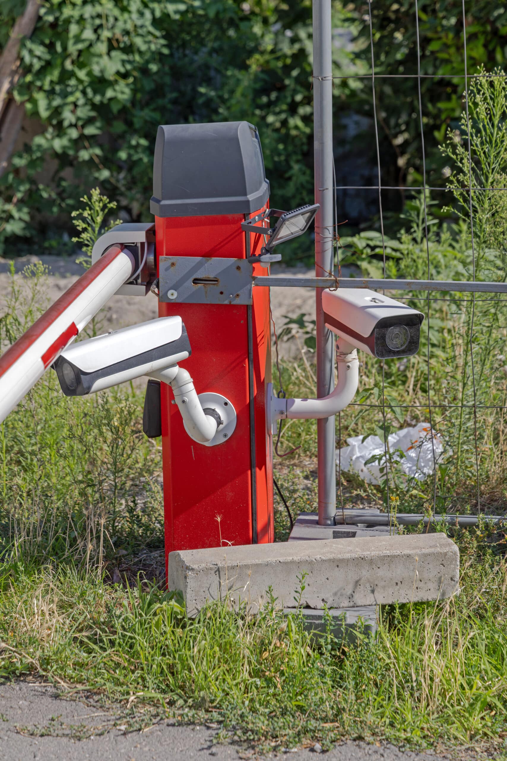 Barrière de sécurité rouge avec deux caméras de vidéosurveillance et un projecteur LED, installée près d'une clôture temporaire et de végétation.