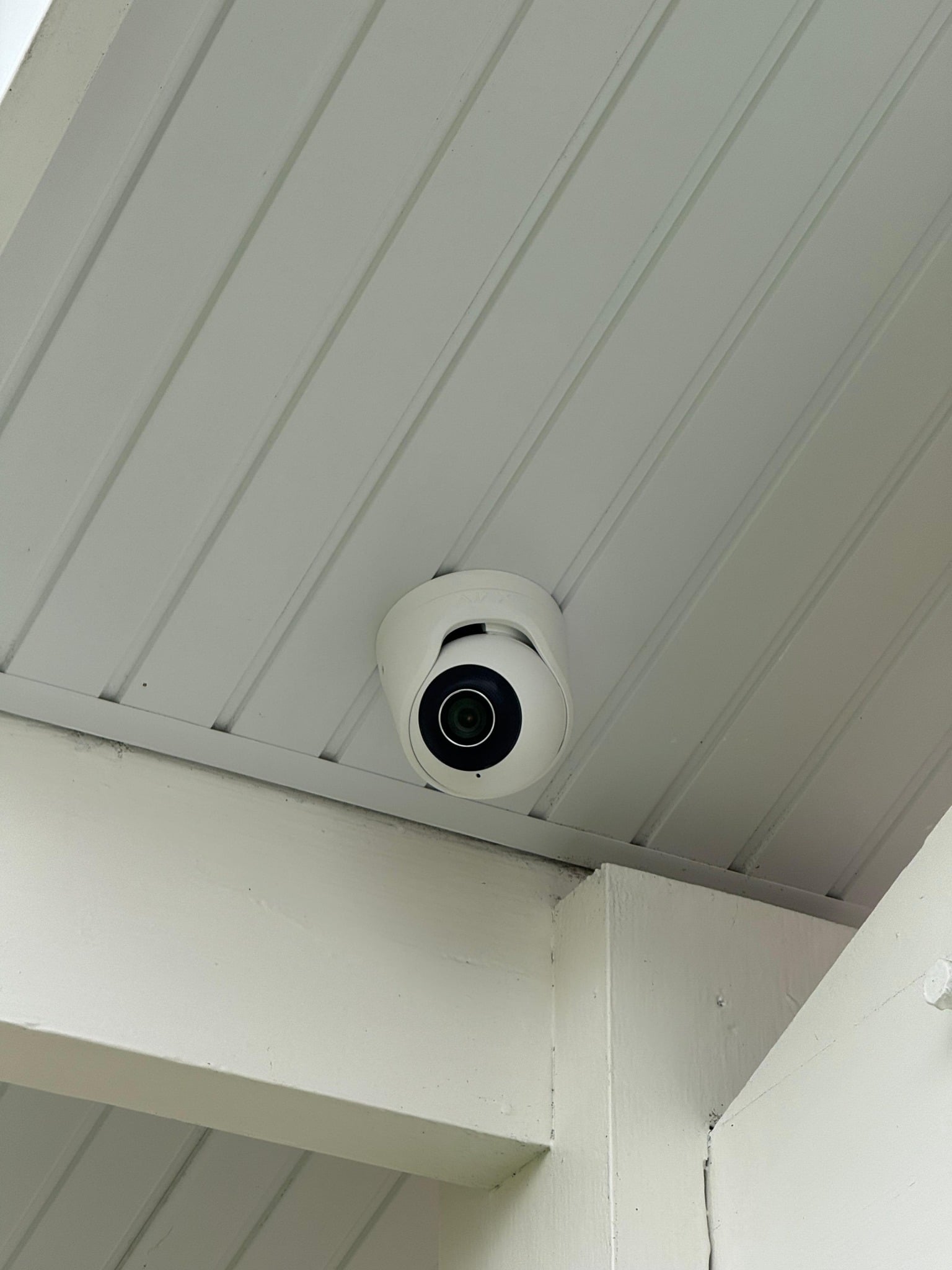 Caméra de surveillance dôme blanche fixée au plafond extérieur à lattes, sous un avant-toit blanc.