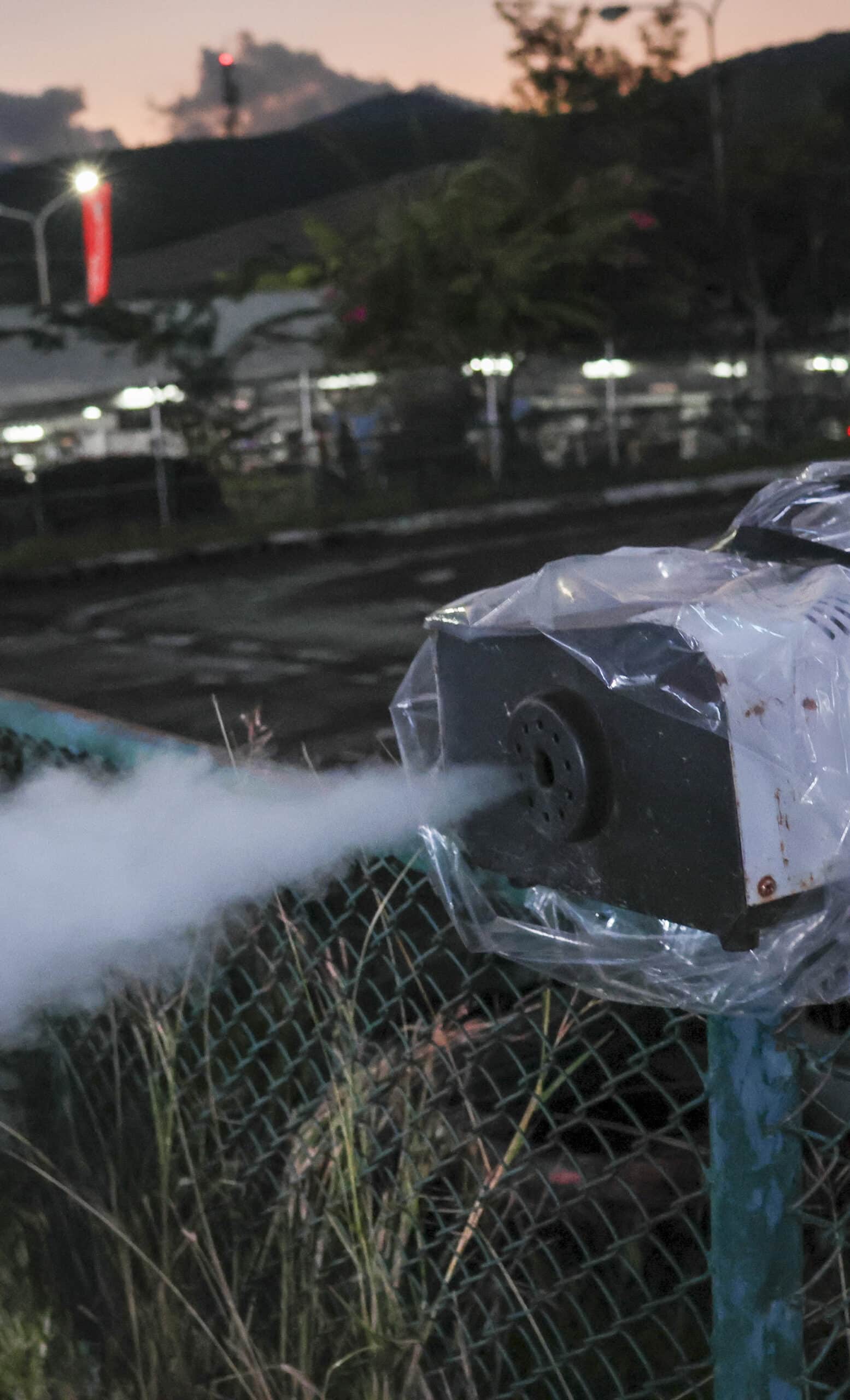 Machine à fumée/brouillard emballée dans du plastique, projetant un jet de fumée blanche à travers une clôture grillagée au crépuscule.
