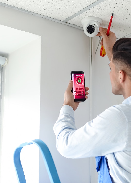 Installation Caméra Sécurité et Maison Intelligente Technicien installant une caméra de sécurité dôme au plafond tout en gérant le système de maison intelligente avec un smartphone affichant un code de verrouillage.