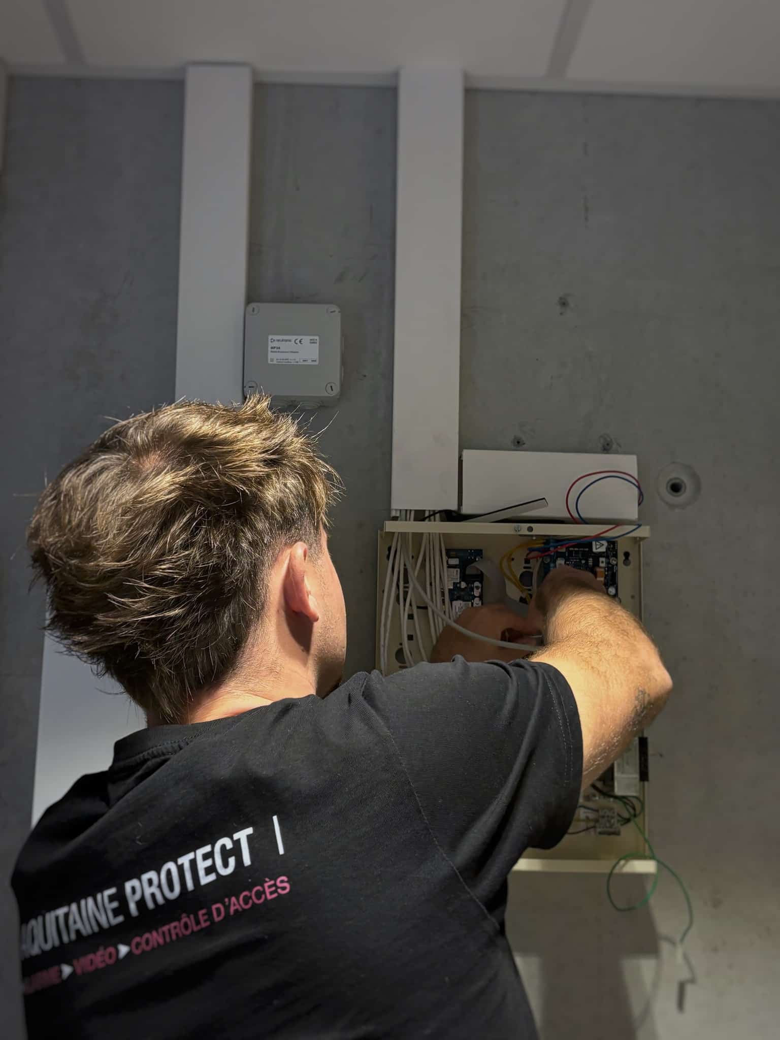 Technicien portant un t-shirt "AQUITAINE PROTECT | ALARME VIDÉO CONTRÔLE D'ACCÈS" câblant un panneau de contrôle électronique mural.