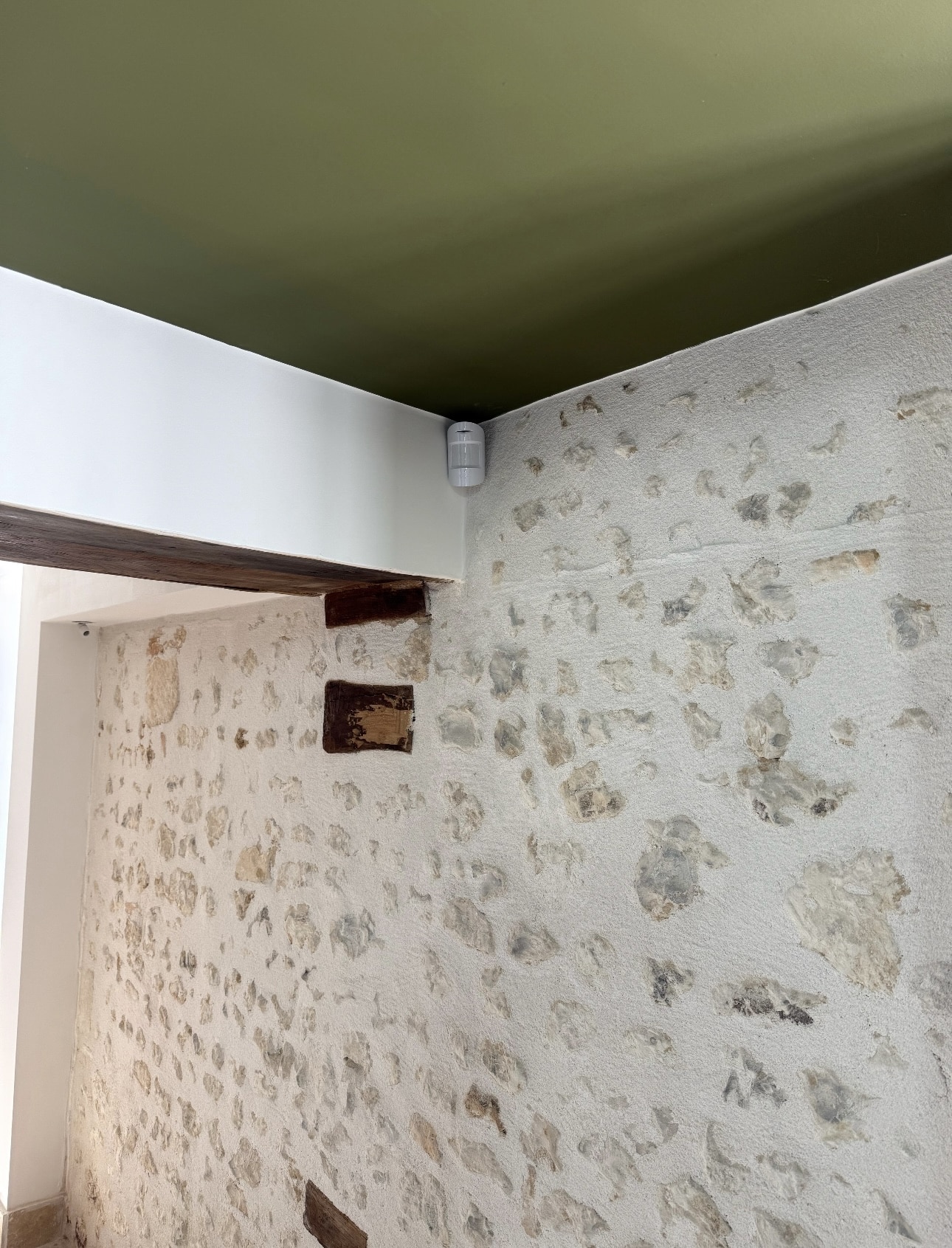 Détail d'un coin intérieur avec mur en pierres apparentes blanchies, poutre en bois sombre, plafond vert olive et détecteur de mouvement blanc.