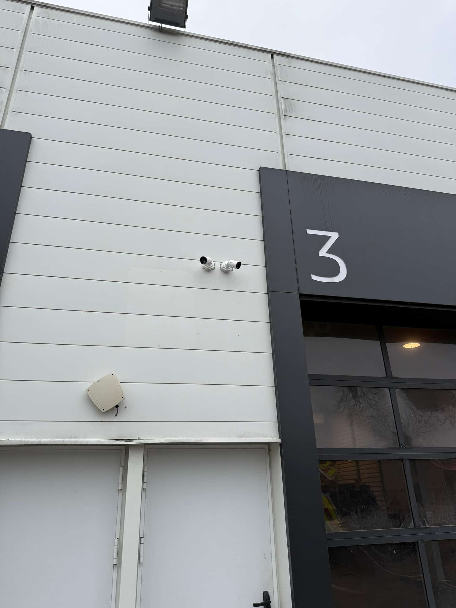 Extérieur d'un bâtiment industriel moderne avec bardage blanc, porte double blanche, et une grande porte vitrée noire marquée du numéro blanc "3". Deux caméras de surveillance sont visibles au-dessus d'un boîtier électrique beige.