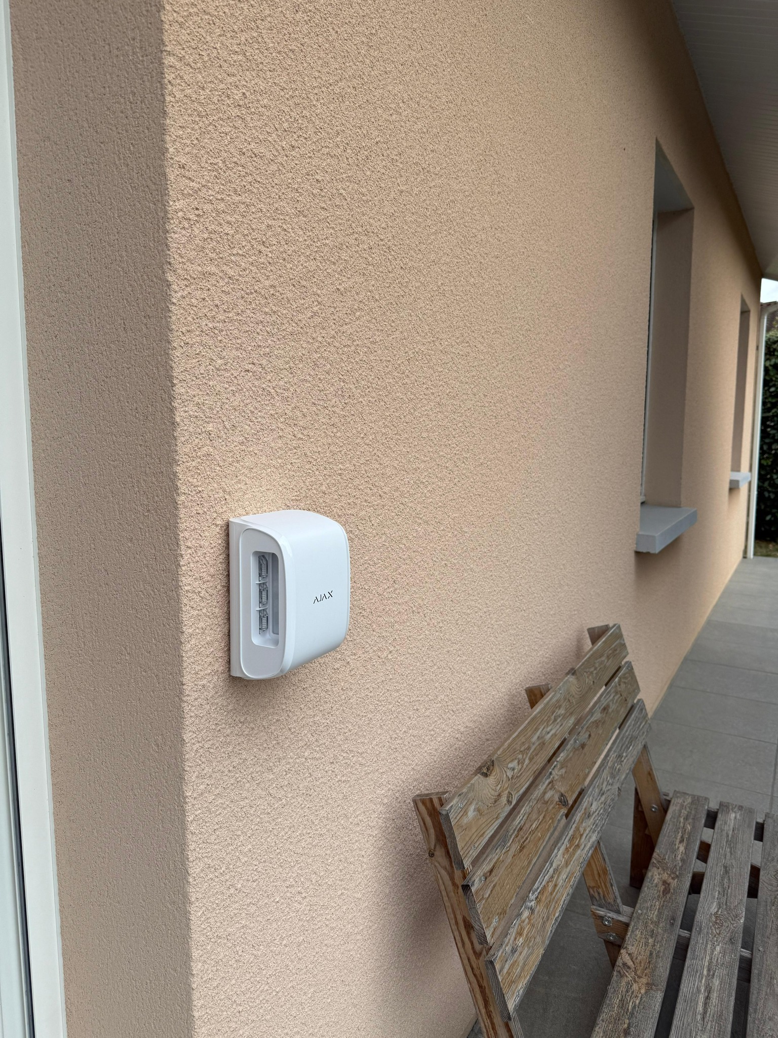 Sirène extérieure Ajax blanche fixée sur un mur crépi beige à côté d'une porte et d'un banc en bois usé.