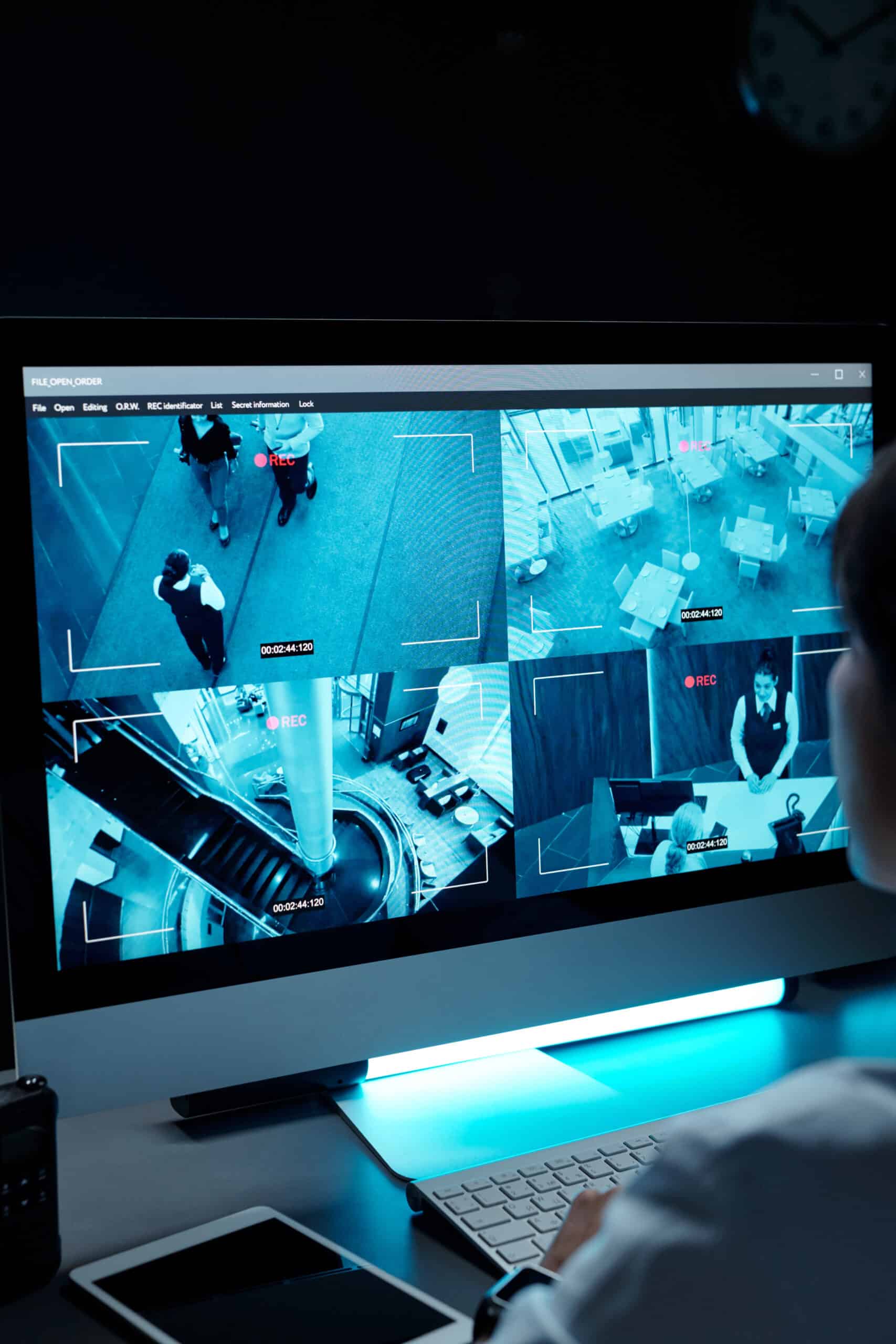 Agent de sécurité surveillant un moniteur affichant quatre flux vidéo de surveillance bleutés, dont un hall d'hôtel et un bureau d'accueil. Le logiciel indique "FILE OPEN ORDER" et "REC".