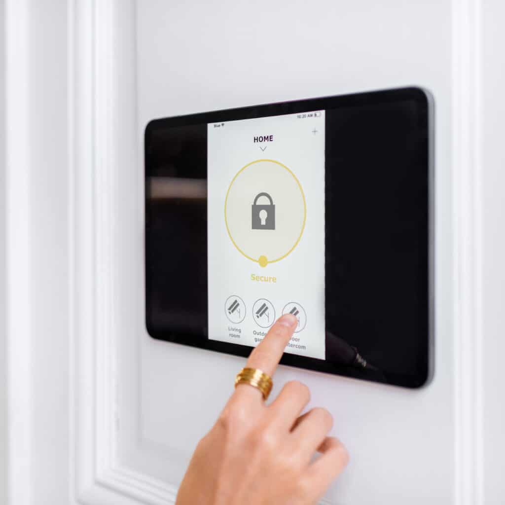 Main avec bague dorée touchant l'icône "Door Intercom" sur un panneau de contrôle domestique "HOME" affichant "Secure".